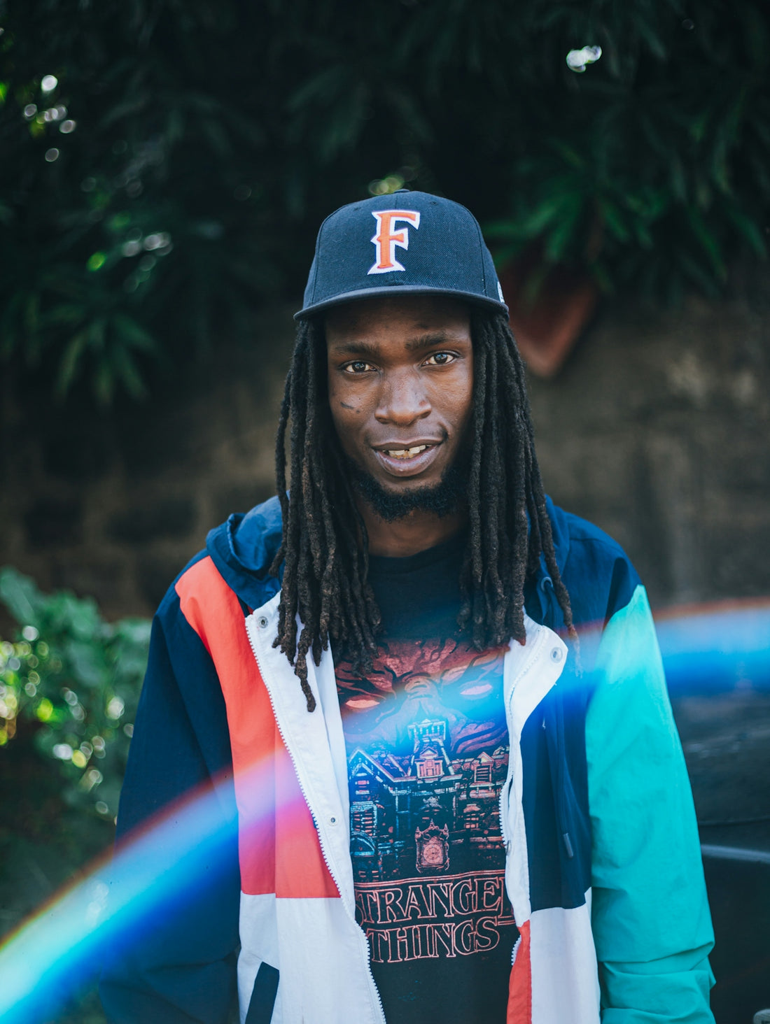 Man with dreadlocks wearing a baseball cap and colorful jacket.
