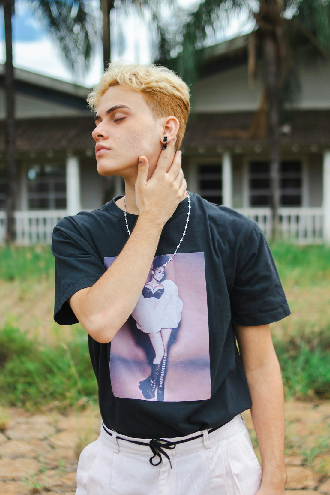 Blonde-haired person touching neck outdoors near a house.